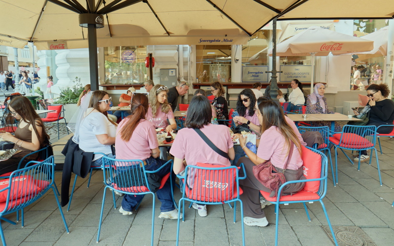 A lively outdoor cafe scene with people enjoying drinks and food under large branded umbrellas